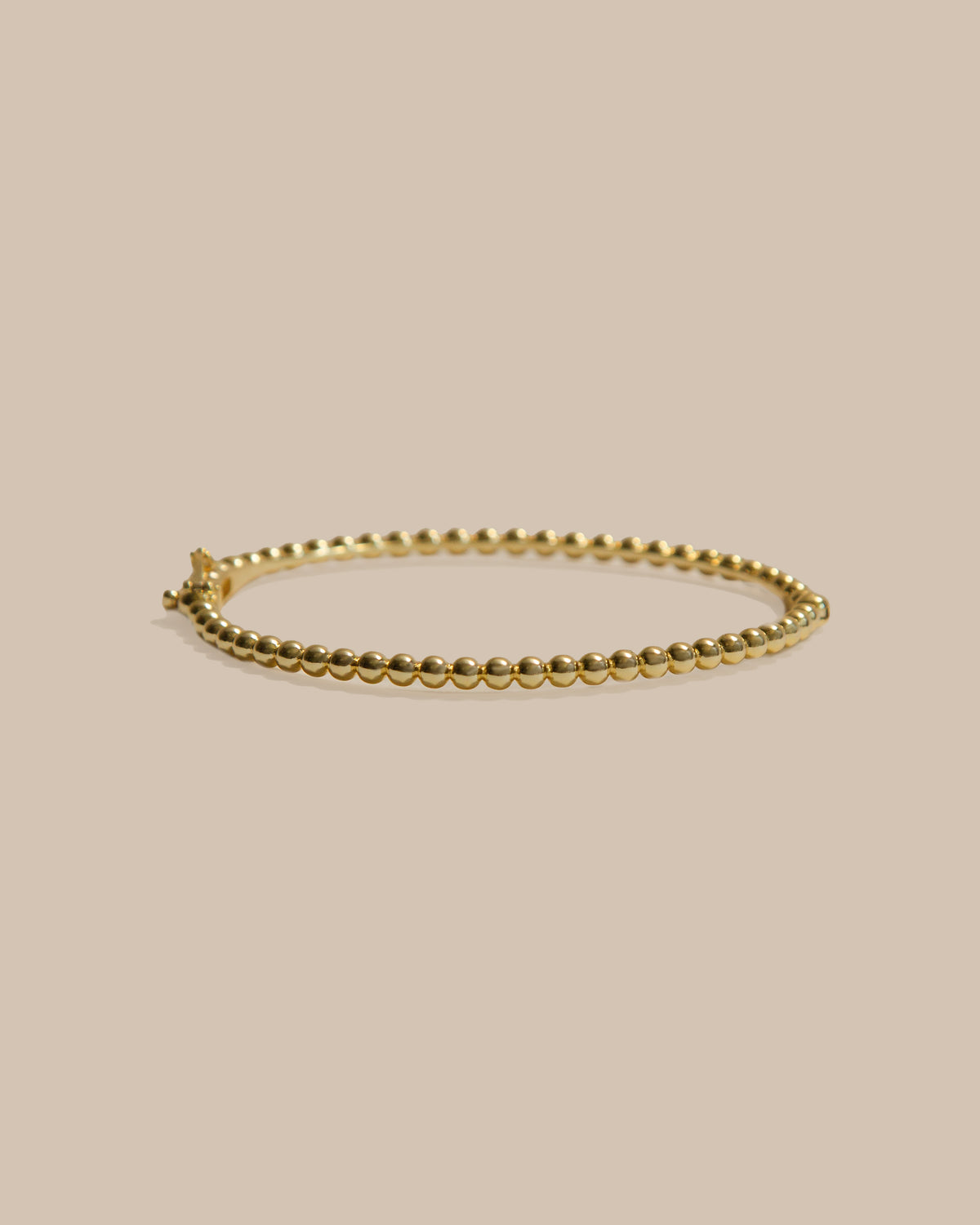 A delicate gold beaded bracelet with a small decorative clasp, displayed on a solid beige background.