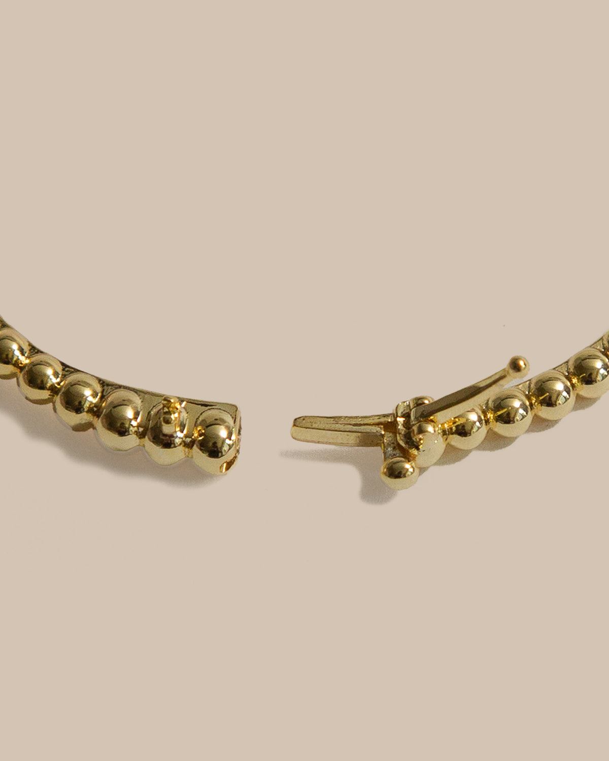 Gold beaded hoop earring shown in a close-up detail shot, highlighting the polished spherical beads and the spring-ring clasp mechanism on a solid beige background.