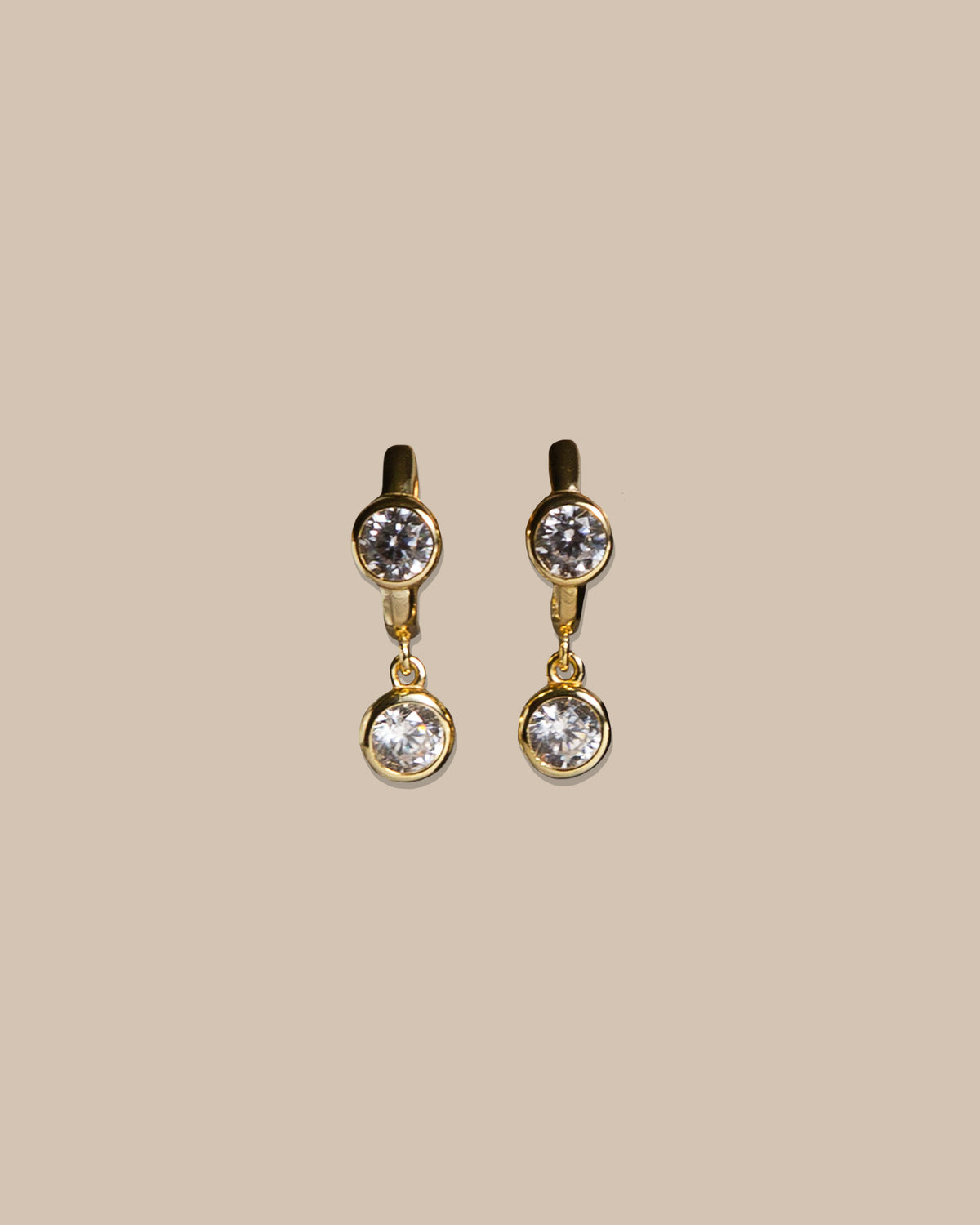 A pair of gold drop earrings with round clear gemstones, displayed against a solid beige background.