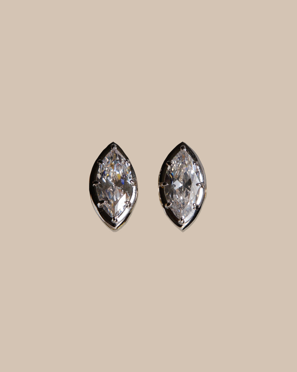 A pair of marquise-cut stud earrings with clear gemstones in a silver-toned setting, centered on a solid beige background.