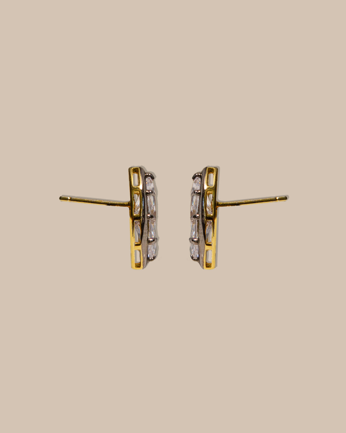 A pair of gold and silver geometric stud earrings with clear baguette-cut stones, shown against a solid beige background.
