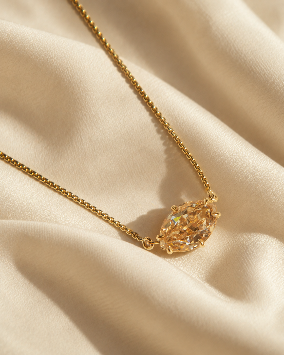 A gold chain necklace with a pear-cut iridescent crystal pendant, displayed on draped cream-colored satin fabric.