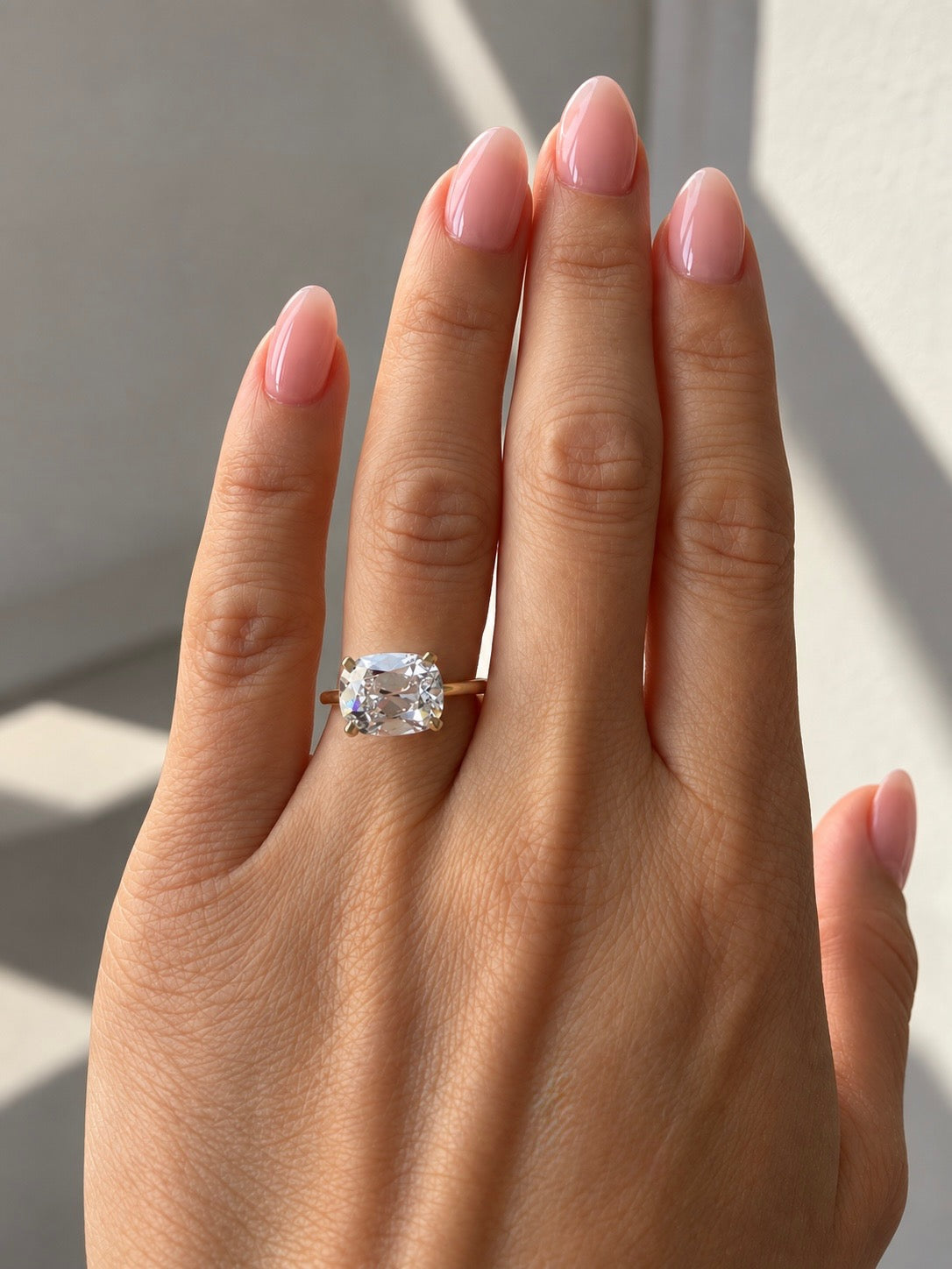 A close-up of a hand with pink manicured nails wearing a gold ring with a cushion-cut clear gemstone, set against a softly lit neutral background.