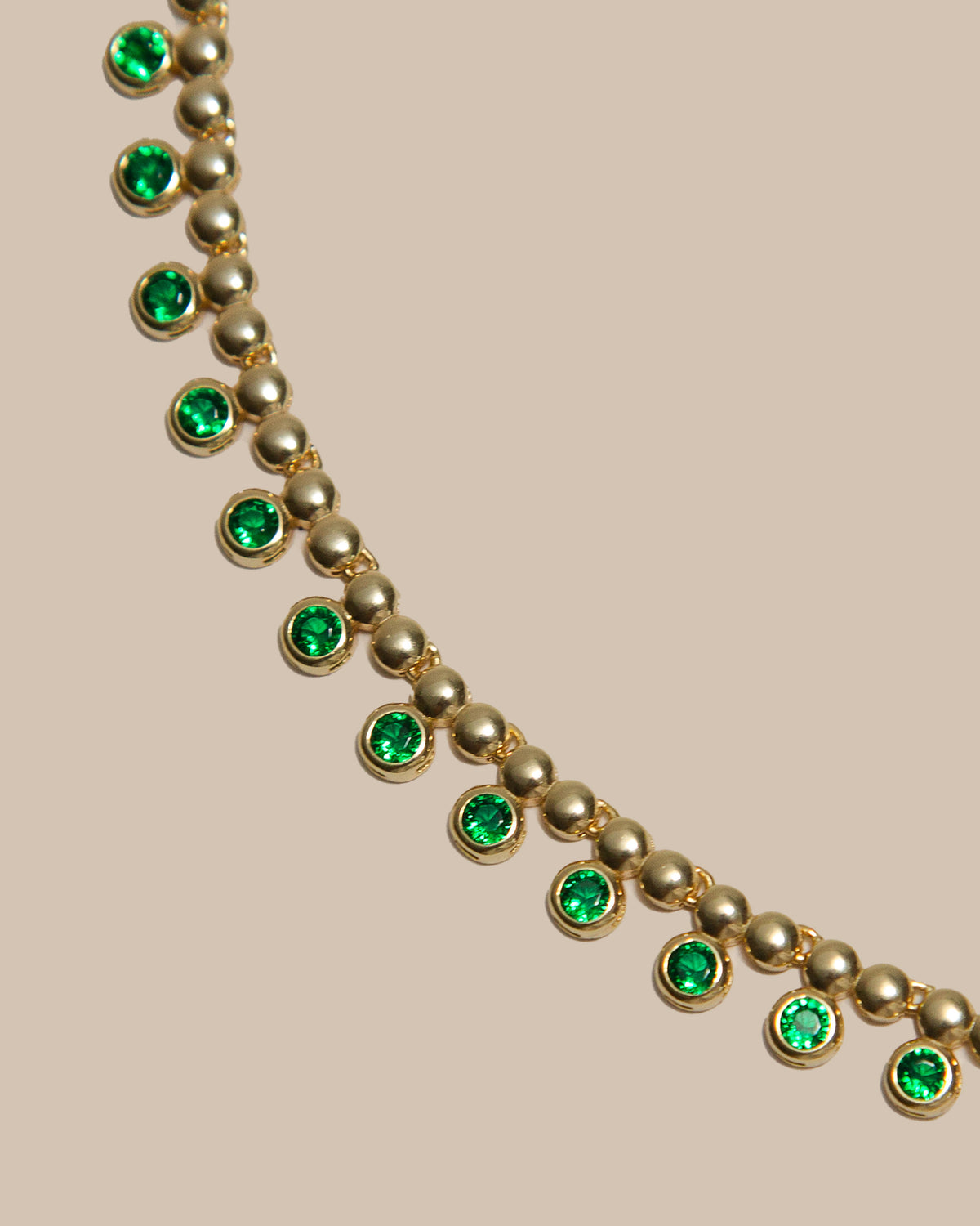 A gold-toned beaded necklace featuring alternating round green gemstone accents, displayed diagonally against a solid beige background.