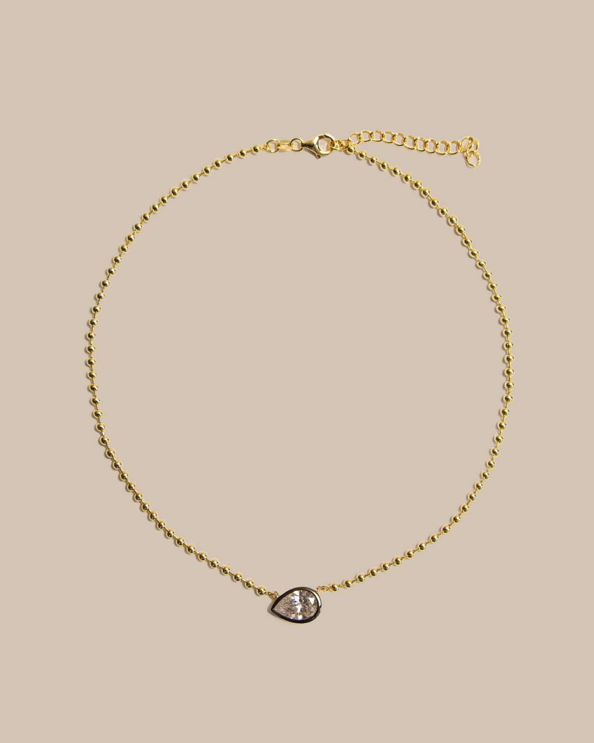 A delicate gold chain necklace with a small teardrop-shaped pendant featuring a clear gemstone, displayed on a solid beige background.