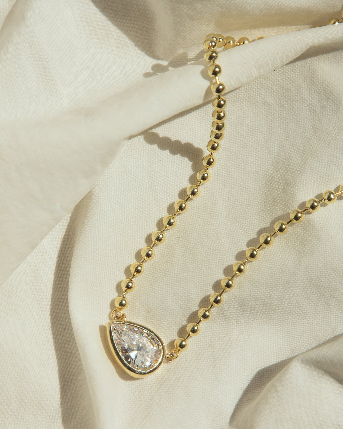 A gold beaded necklace with a teardrop-shaped clear gemstone pendant, styled on draped cream-colored fabric.