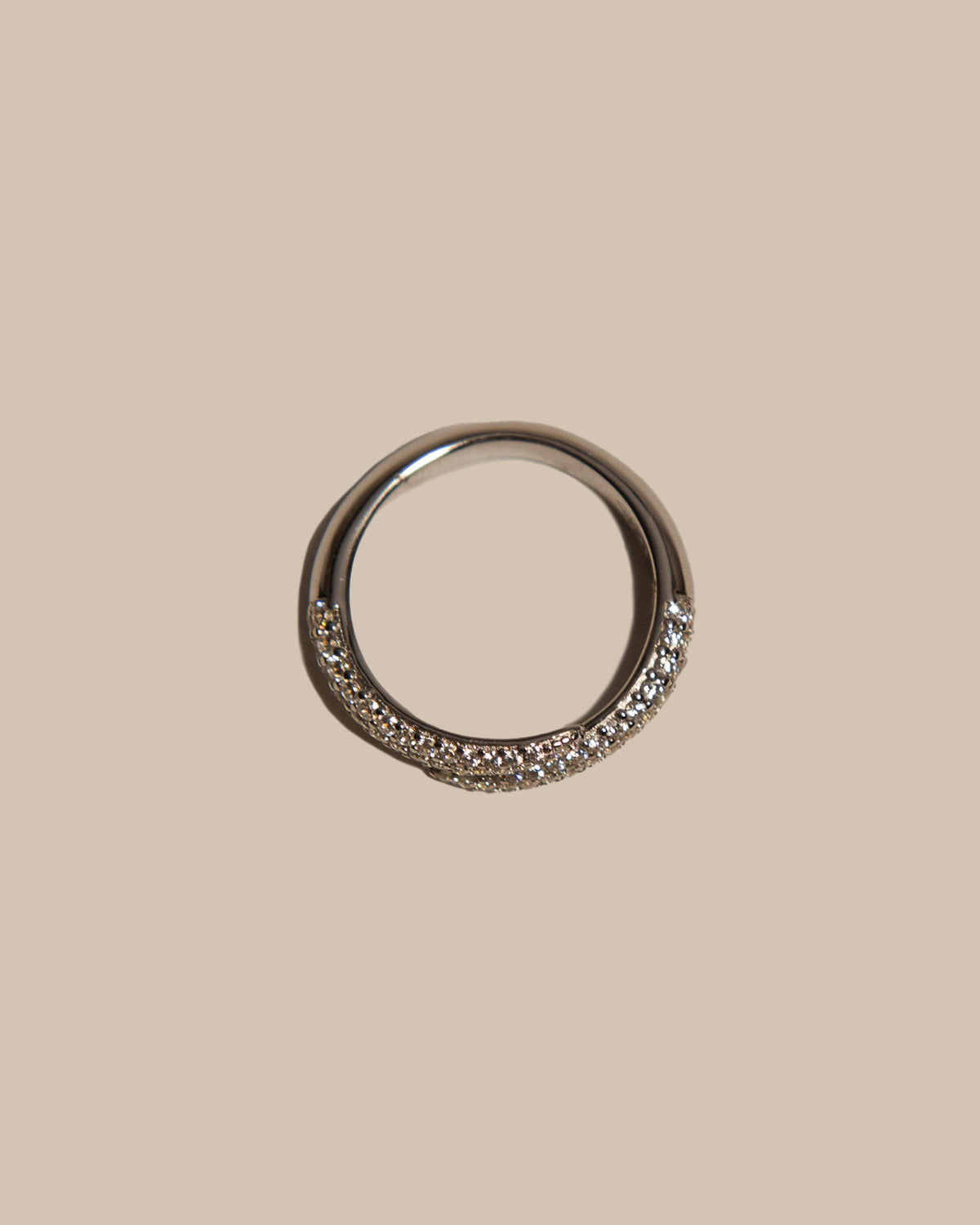 A silver-toned ring with a row of clear pavé-set stones along the front half, shown against a solid beige background.