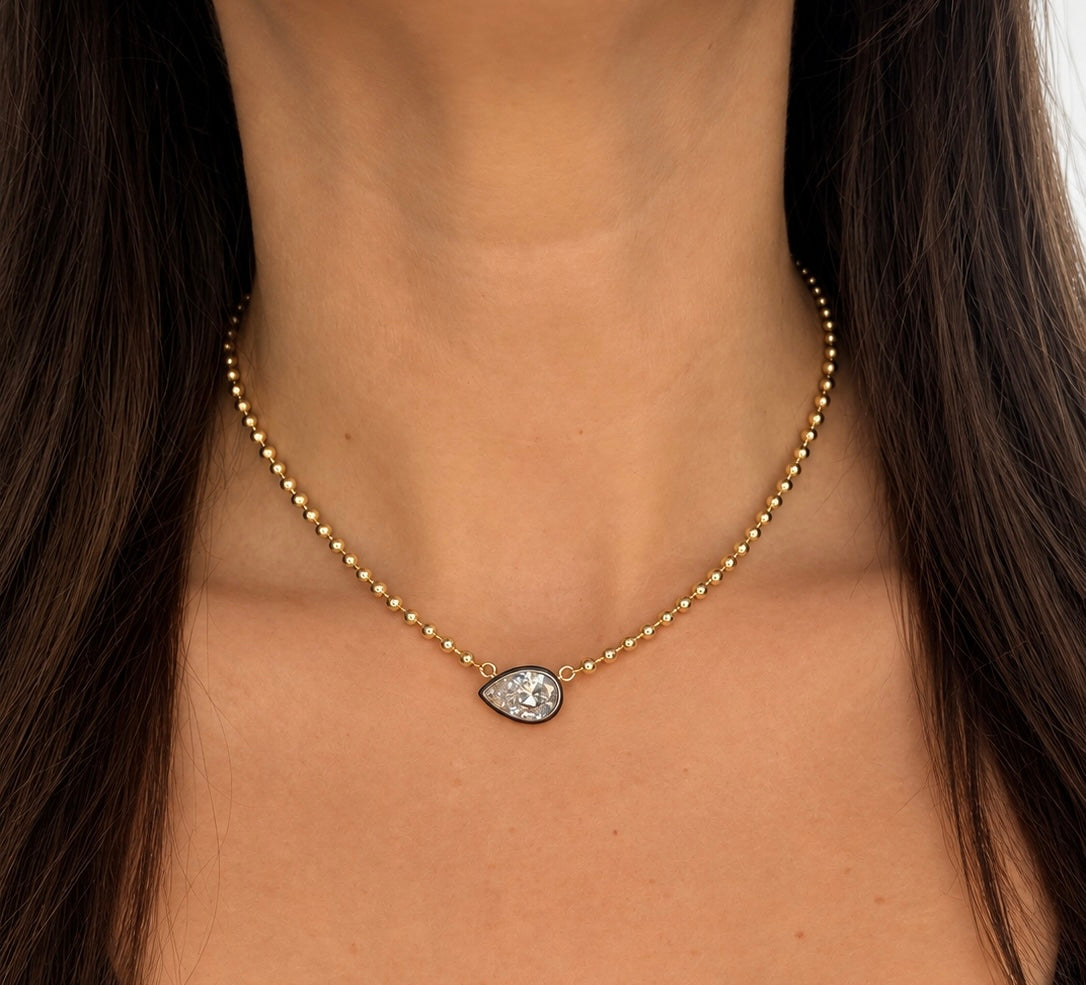 A close-up of a model's neck wearing a delicate gold beaded necklace with a teardrop-shaped clear crystal pendant.