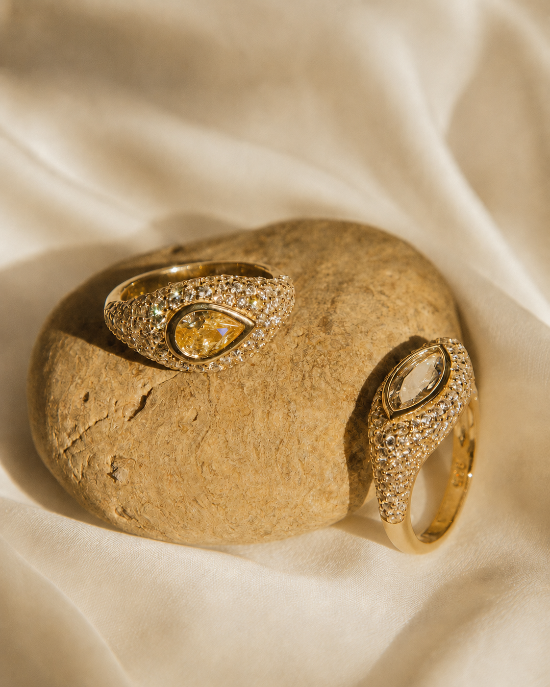 Two gold rings with pavé-set clear stones and teardrop-shaped yellow gemstones, displayed on a smooth beige stone over draped cream fabric.