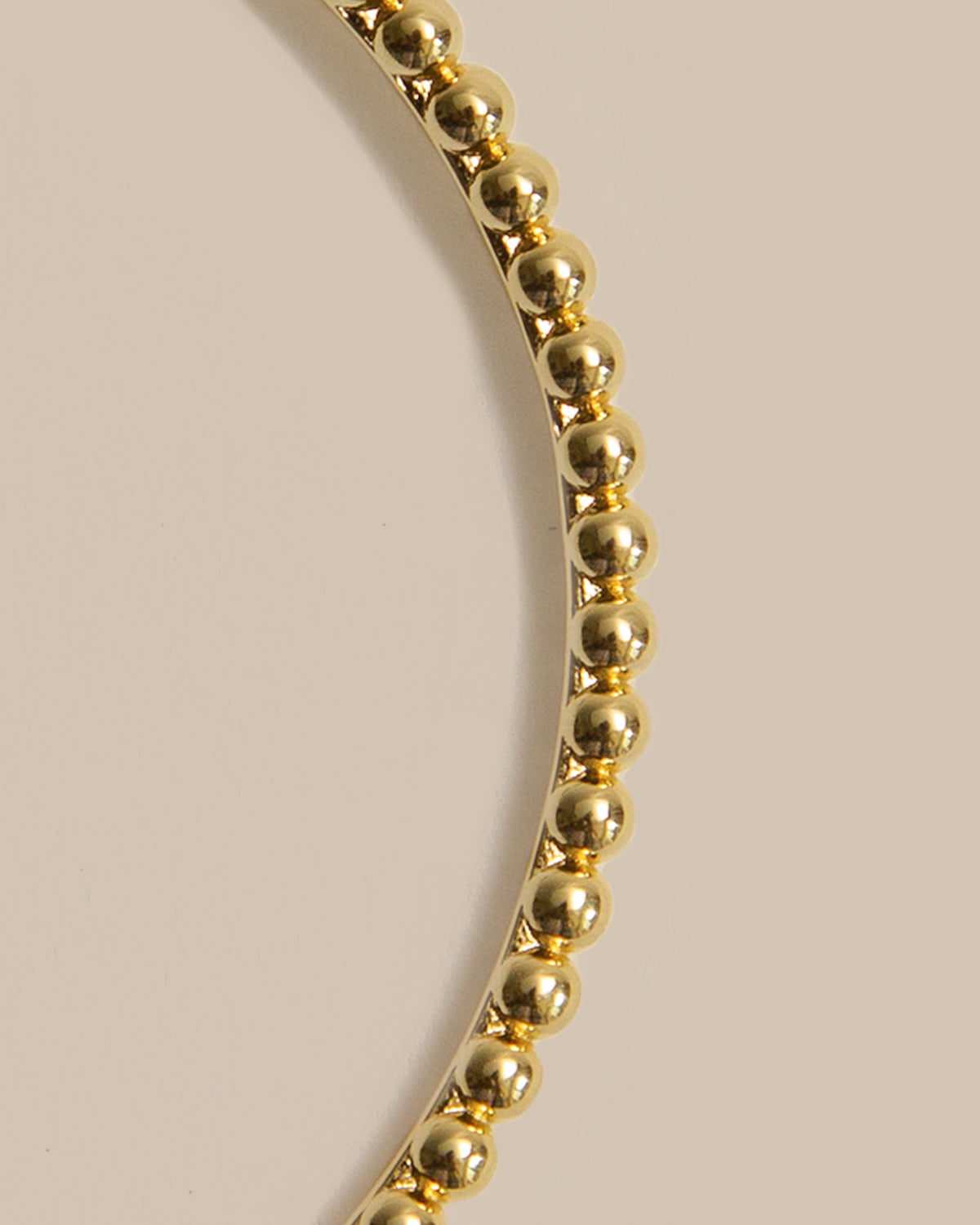 A close-up of a gold beaded bracelet or bangle shown in a curved shape against a solid beige background.