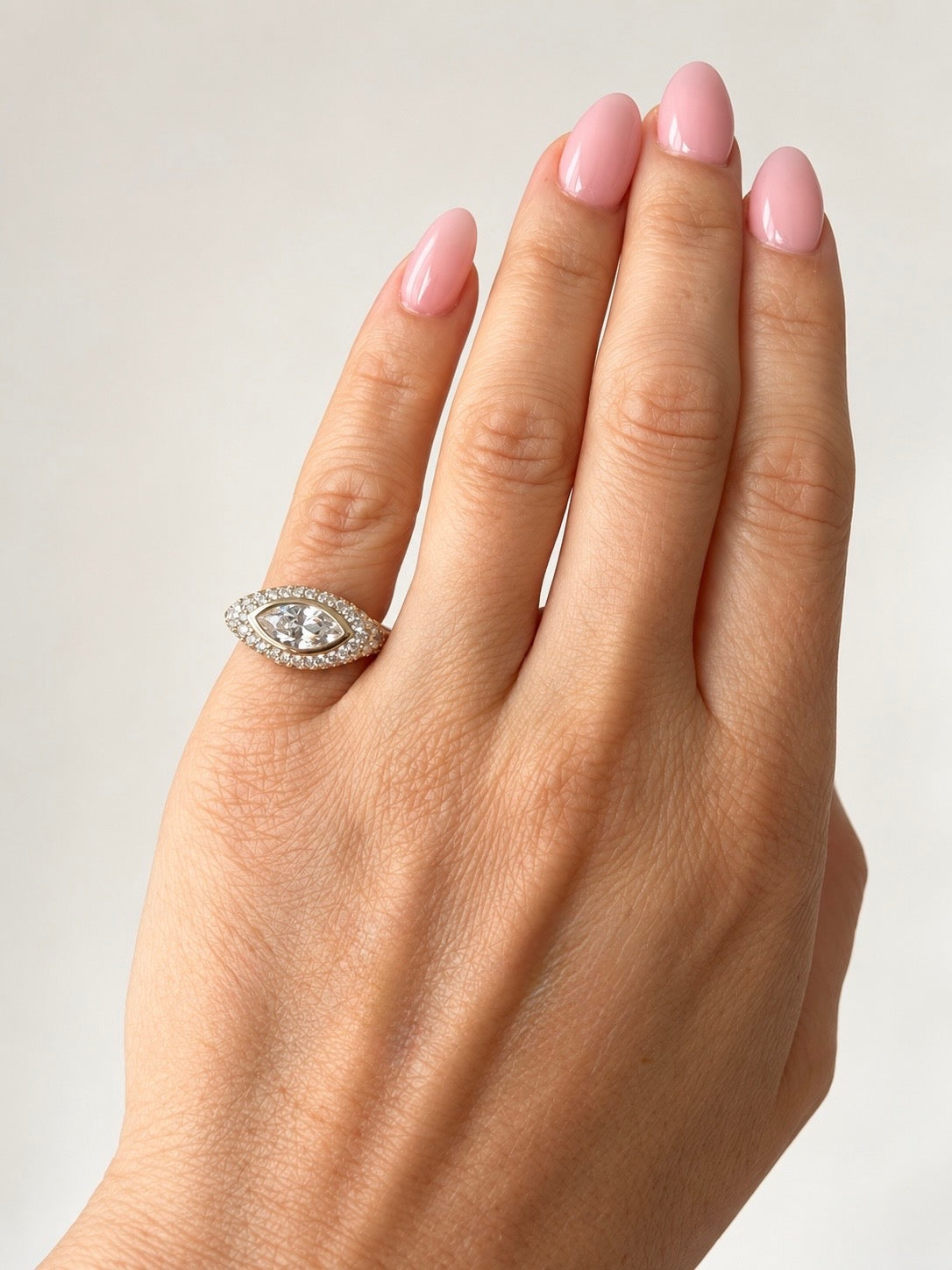 A close-up of a hand with pink manicured nails wearing a silver-toned eye-shaped ring set with small clear stones.