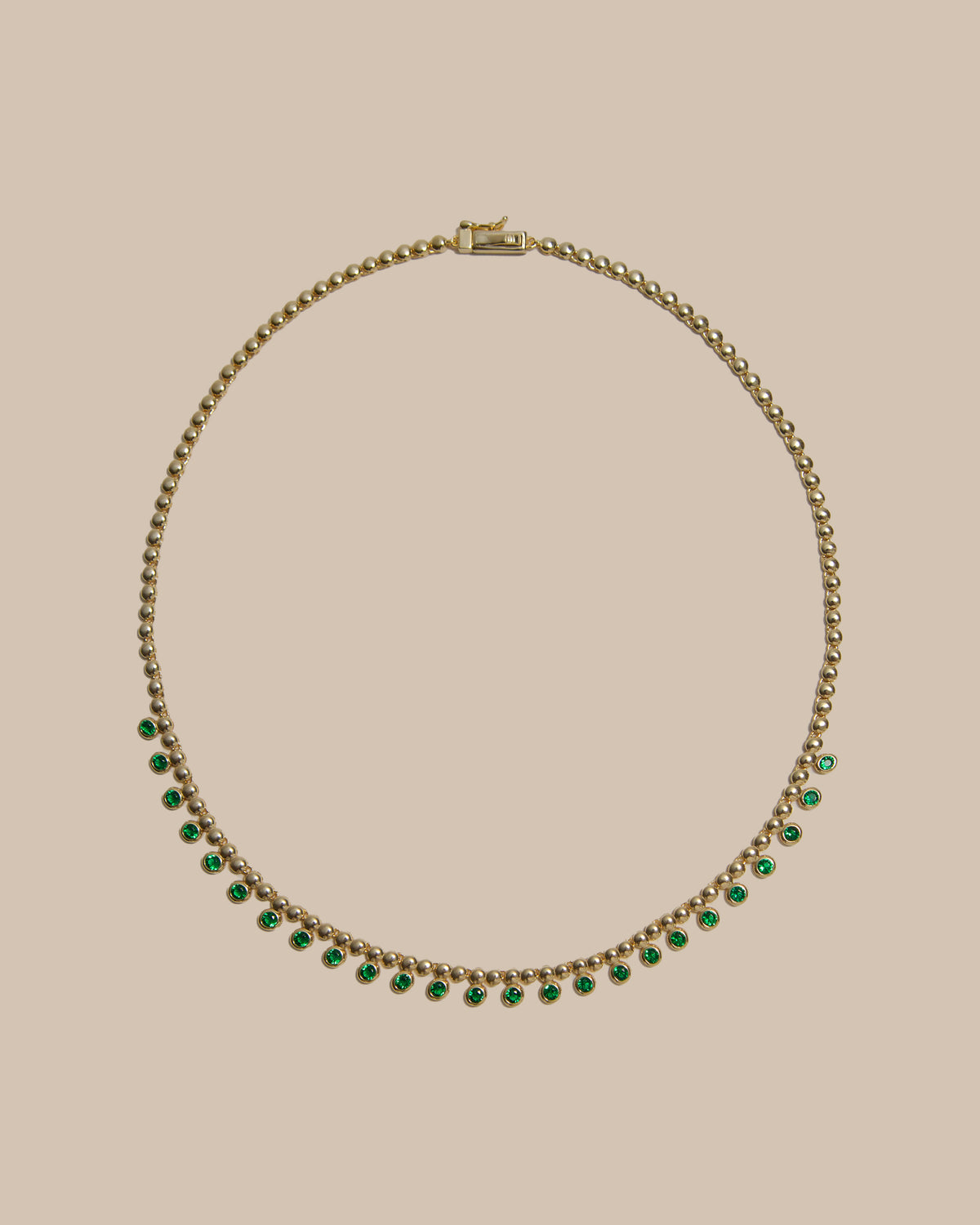 A gold chain necklace featuring a row of small, evenly spaced green gemstones along the lower curve, displayed against a solid beige background.
