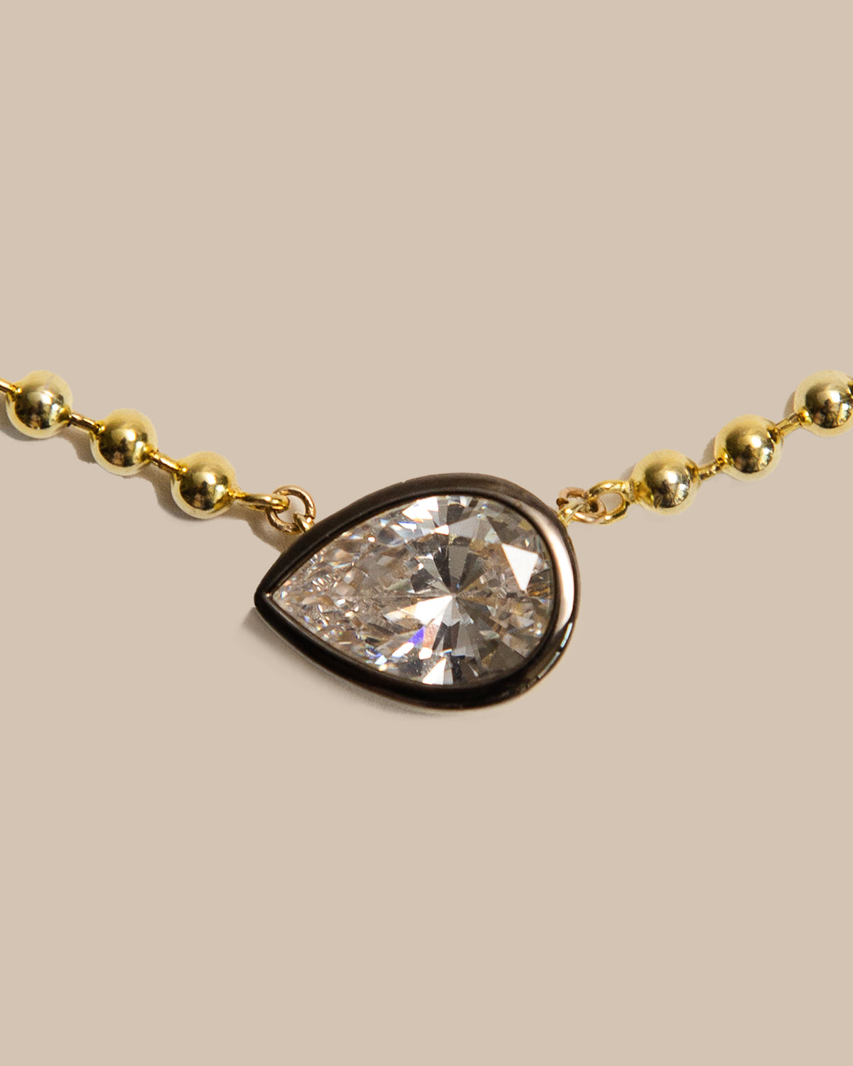A close-up of a gold beaded chain necklace featuring a teardrop-shaped clear gemstone pendant with a polished gold border, set against a solid beige background.