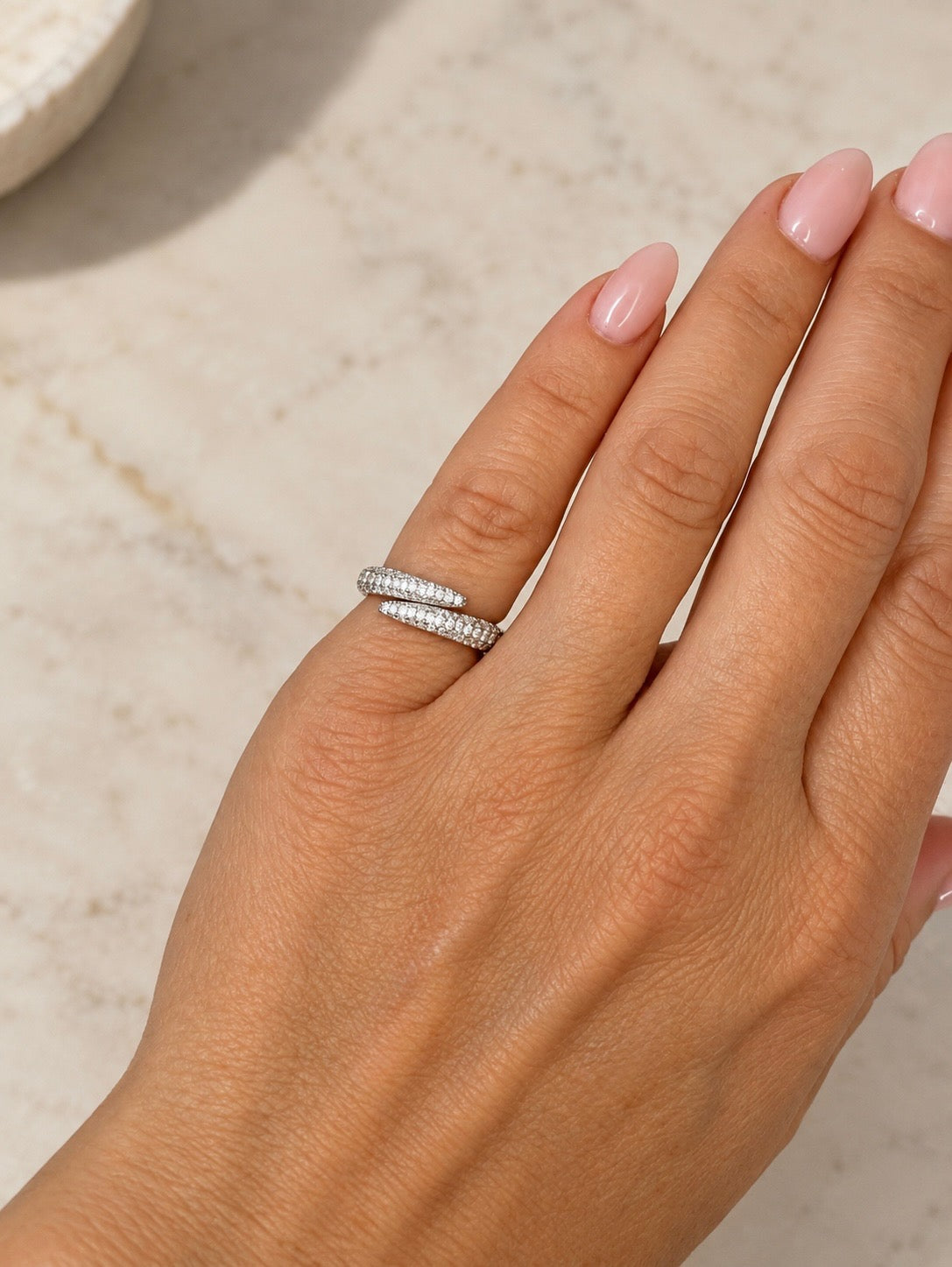 A close-up of a hand wearing a silver band ring set with small clear stones, photographed against a light marble surface.