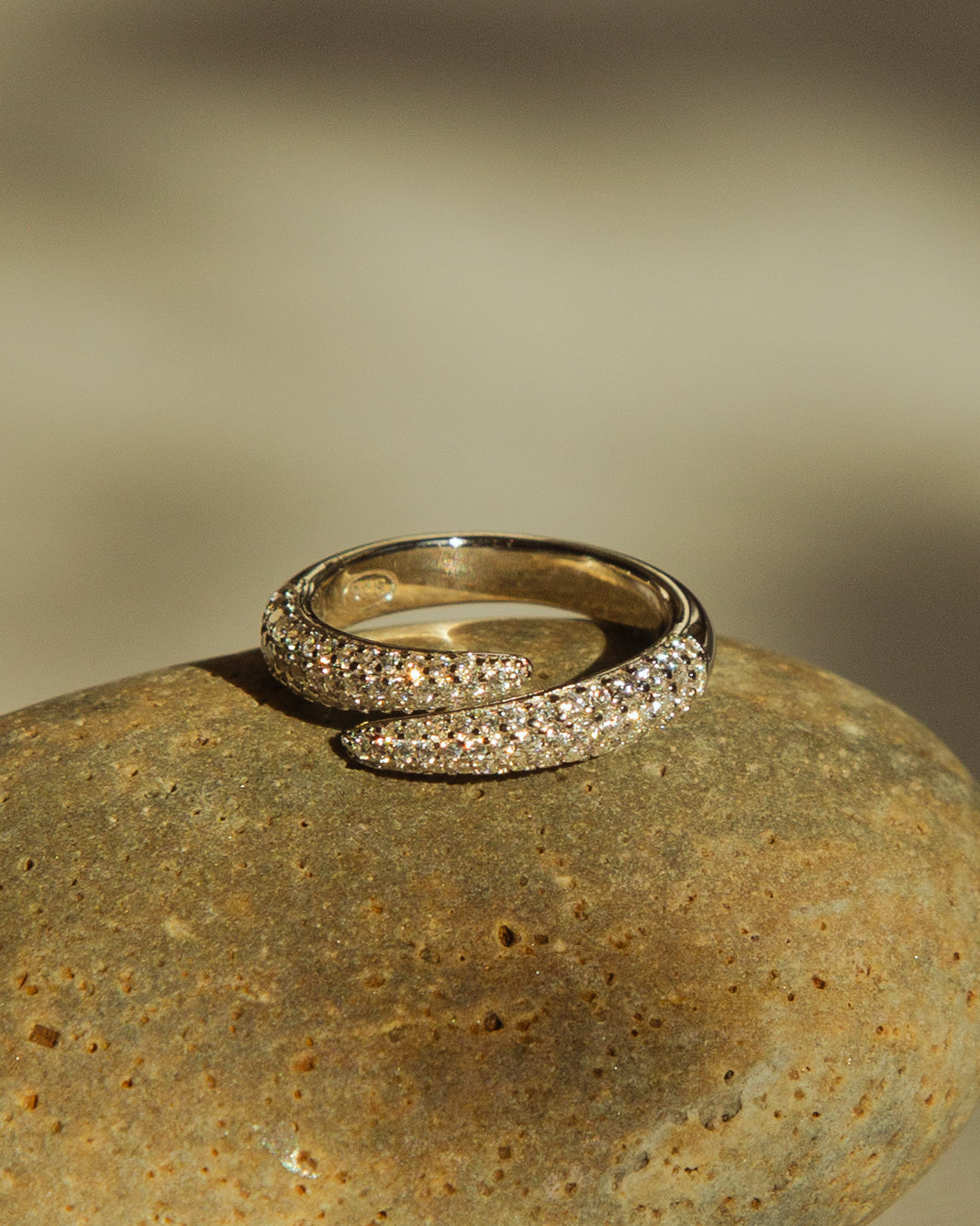 A silver wrap ring with pavé-set clear stones, resting on a textured brown stone with a soft-focus beige background.