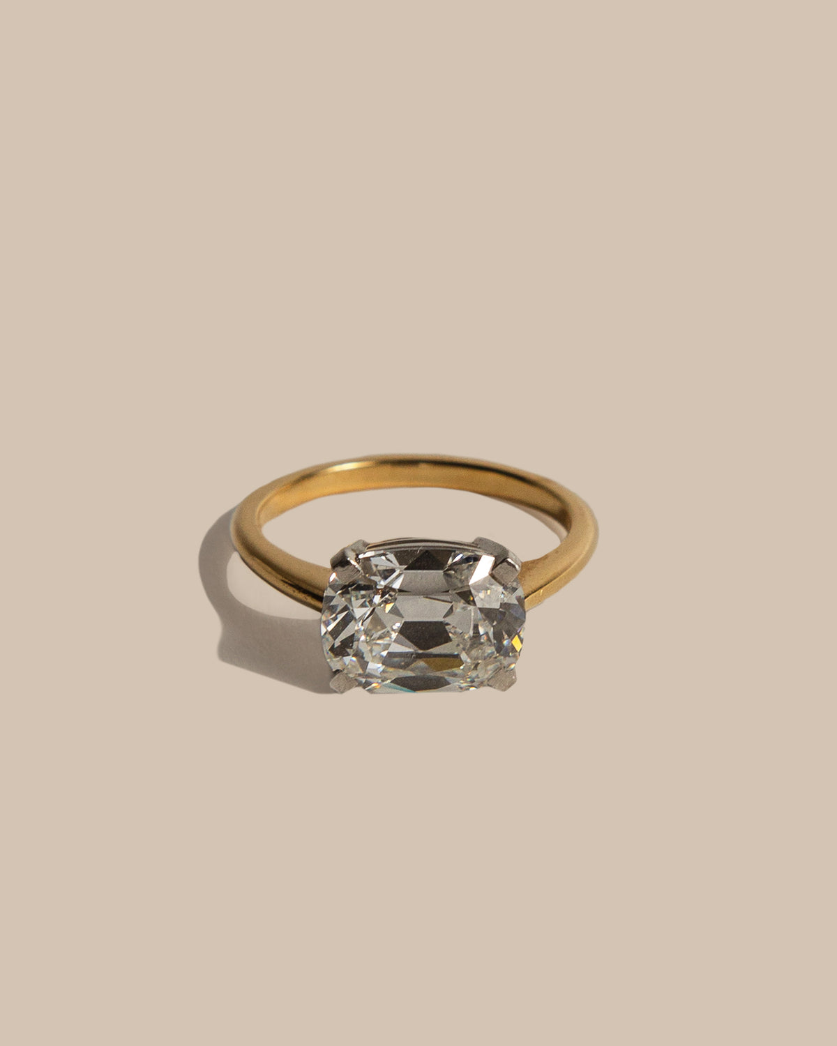 A gold ring with a cushion-cut clear gemstone, photographed on a solid beige background with a soft shadow.