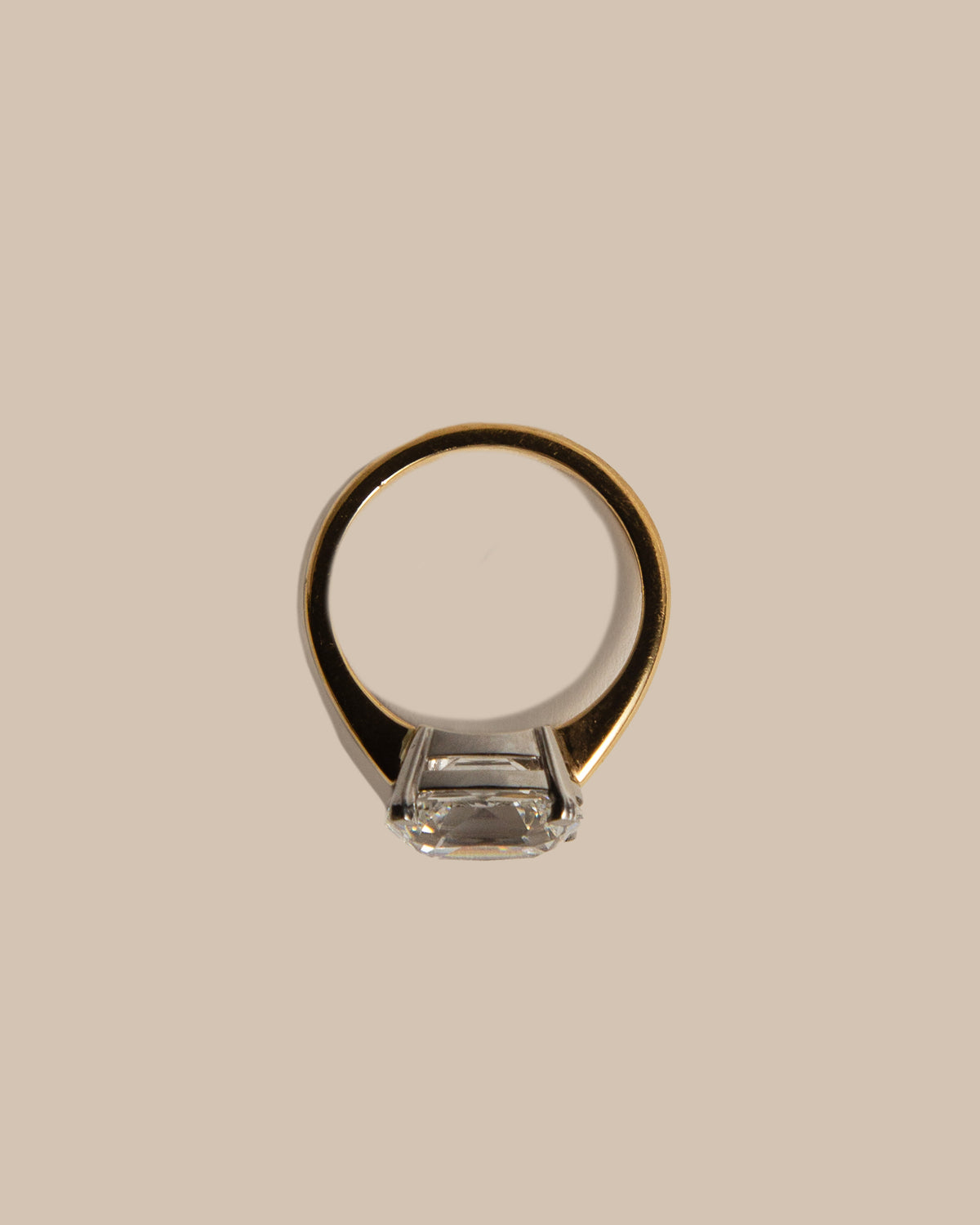 A minimalist gold ring with a clear rectangular gemstone, shown from a top-down view on a solid beige background.