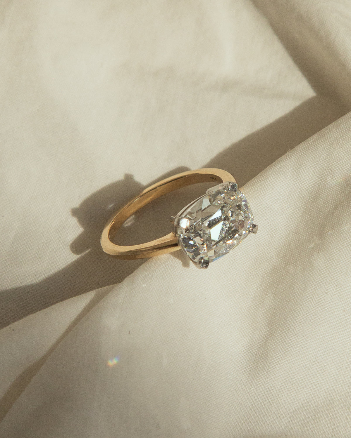 A yellow gold ring with a large clear emerald-cut diamond solitaire, resting on draped cream-colored fabric with a soft shadow.