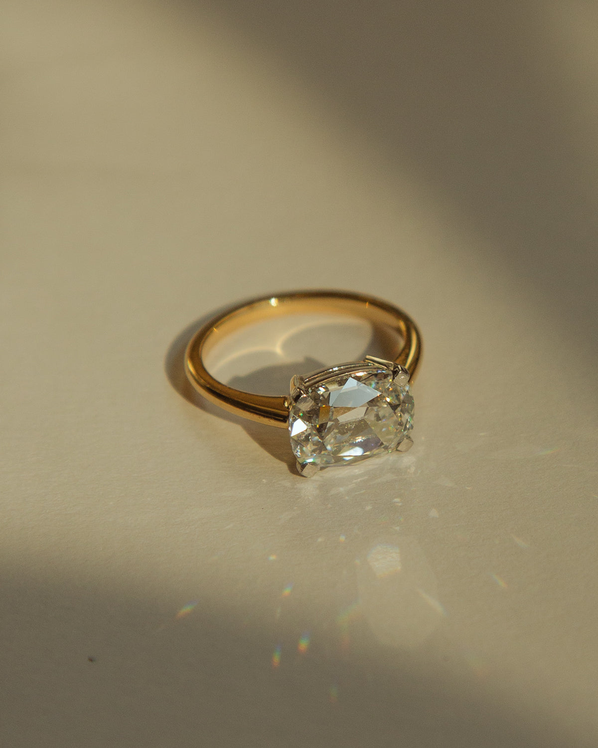 A gold ring with a large cushion-cut clear gemstone, photographed on a warm beige surface with dramatic sunlight and shadows.