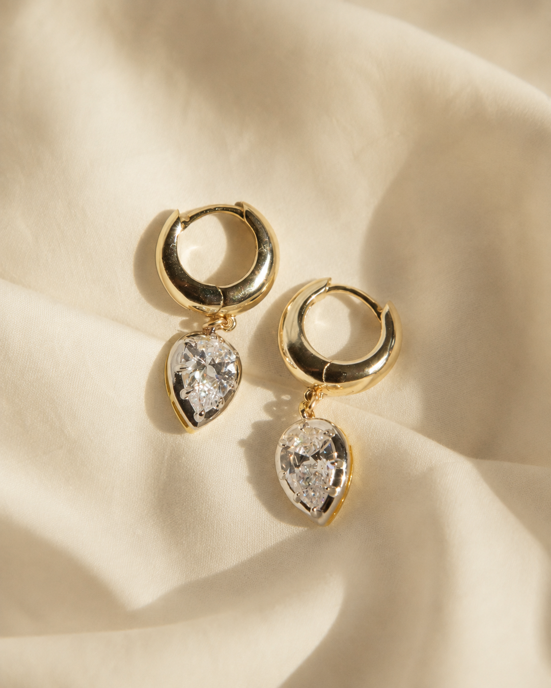 A pair of gold huggie hoop earrings with pear-cut clear gemstones, displayed on a draped cream-colored fabric background.