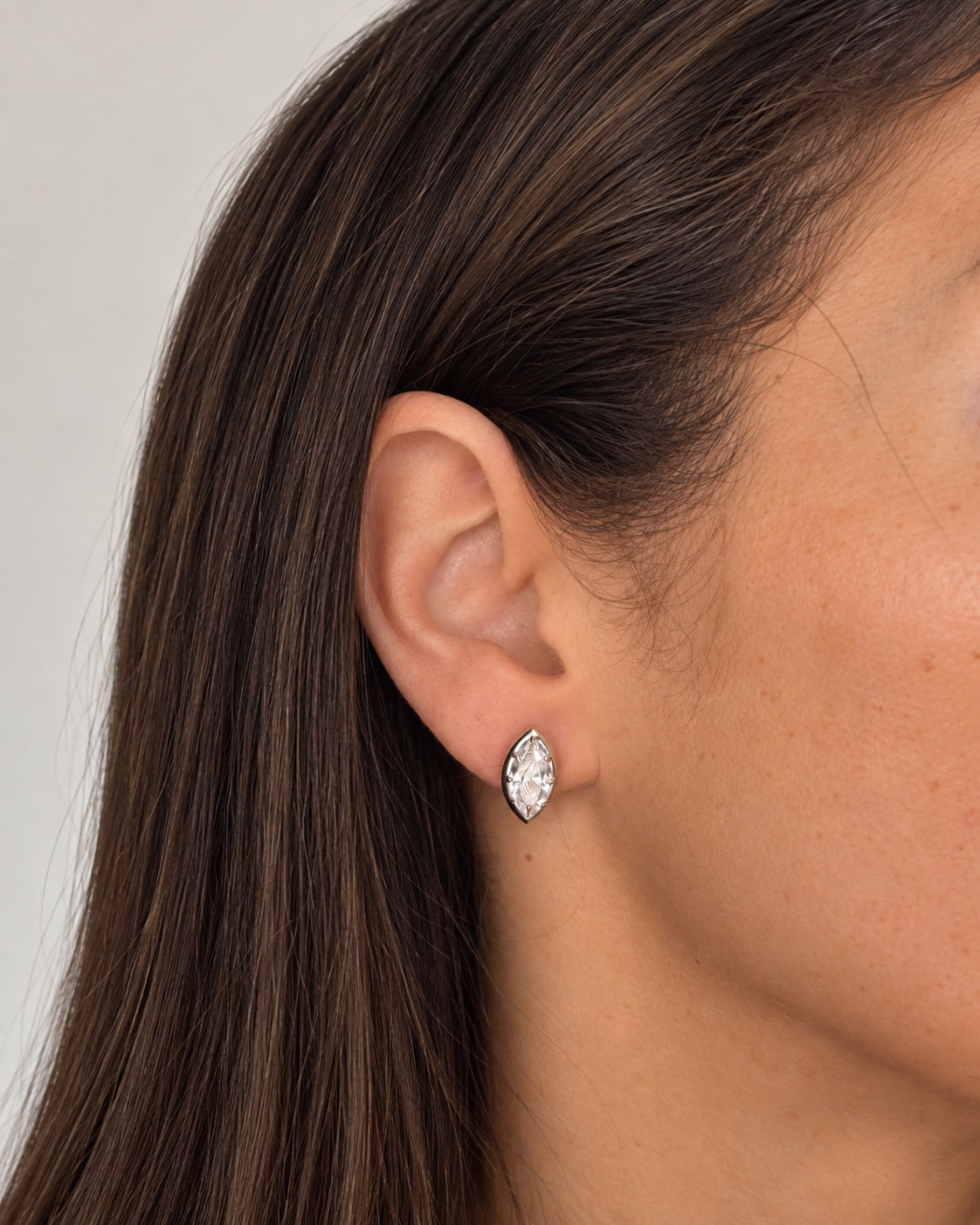 A close-up of a woman's ear wearing a small marquise-shaped stud earring with a clear faceted gemstone in a silver-toned setting.