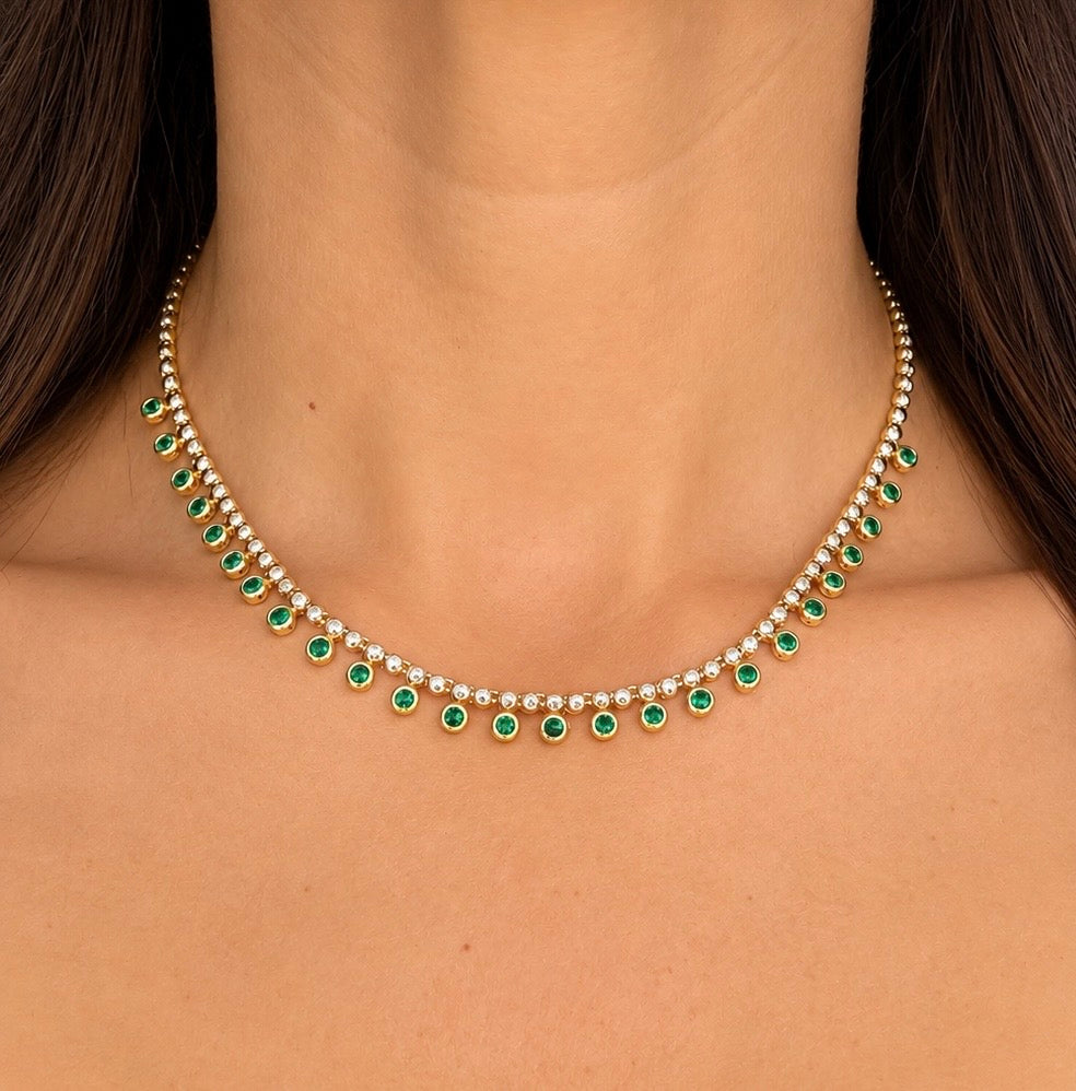 A close-up of a gold necklace with green emerald-cut stones and small clear stones, worn on a model's neck.
